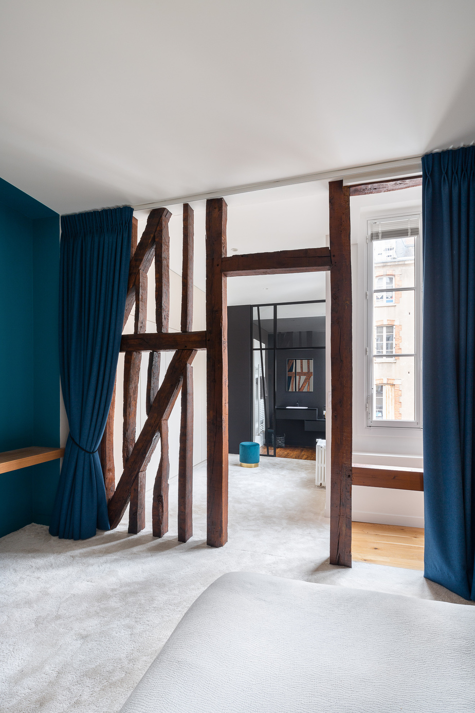 Photographie appartement Rennes : salle de bain parentale vue à travers le colombage, moquette couleur "Mastic" dans la partie dressing, verrière en miroir du colombage en séparation de la salle de bain, meuble vasque noir sur mur noir, moquette dans la partie chambre, rideaux bleutés couleurs des murs devant le colombage pour fermer l'espace nuit