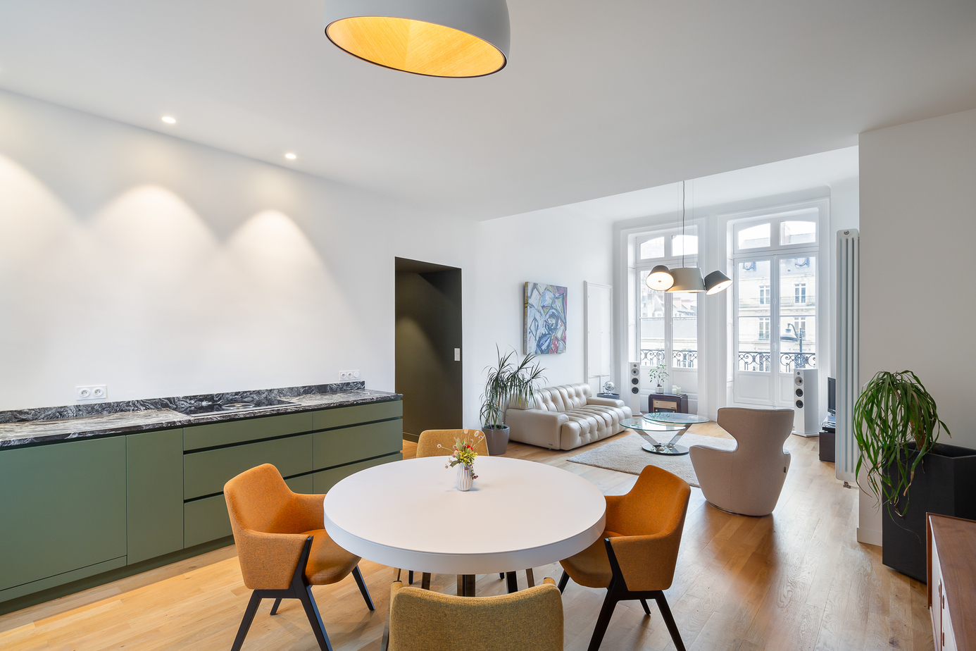 Photographie appartement Rennes : esapce cuisine & salle à manger avec cuisine en agencement laquée Kaki, plan de travail en pierre Cheyenne avec plaque de cuisson Bora, parquet refait à neuf, table à manger ronde blanche et chaises jaunes, suspension Vibia Duo blanche et bois au-dessus, enfilade ancienne chinée / l'ensemble est ouvert sur le salon