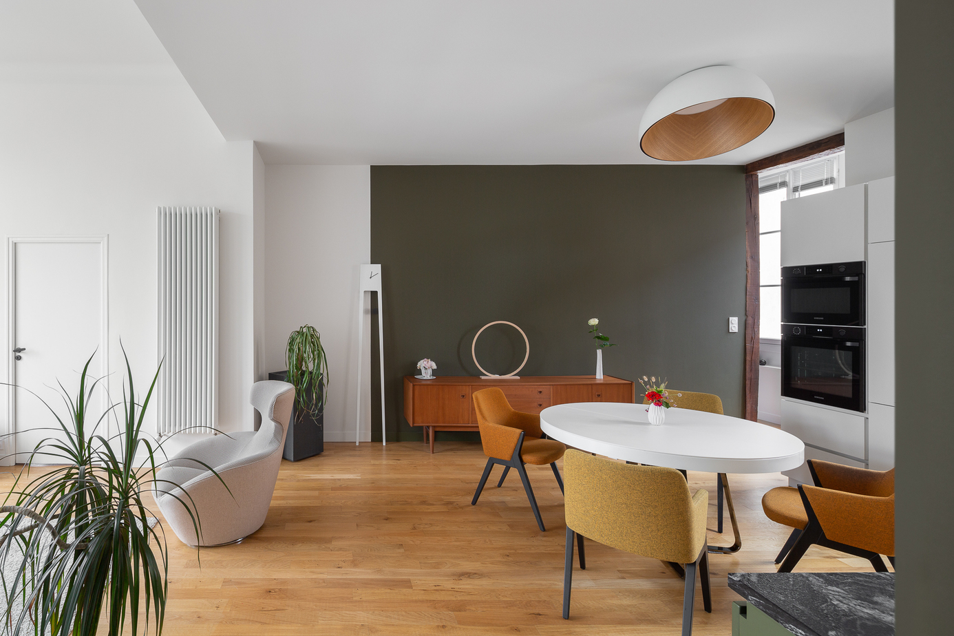 Photographie appartement Rennes : espace cuisine & salle à manger avec cuisine en agencement laquée Kaki, plan de travail en pierre Cheyenne avec plaque de cuisson Bora, parquet refait à neuf, table à manger ronde blanche et chaises jaunes, suspension Vibia Duo blanche et bois au-dessus, enfilade ancienne chiné / l'ensemble est ouvert sur le salon